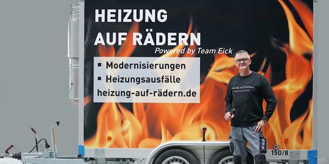 Ein Mann steht neben einem Anhänger mit einem auffälligen, feurigen Design, das Heizungsdienstleistungen bewirbt.